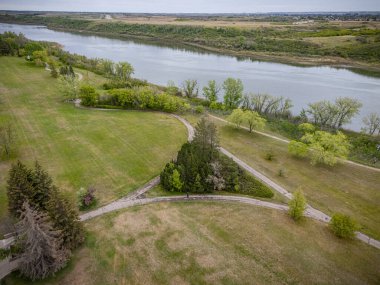 Bir nehir manzarası ve küçük bir gölete giden yolu olan bir park. Park ağaçlar ve çimlerle çevrili ve etrafta dolaşan birkaç kişi var.