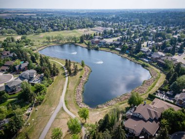 Arka planda evler olan bir göl manzarası. Göl ağaçlarla çevrili ve ona giden bir yol var.