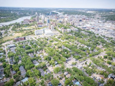Şehir manzarası yukarıdan, içinden bir nehir akıyor. Şehir evler ve binalarla dolu ve caddelerde birçok araba var. Gökyüzü açık ve güneş parlıyor.