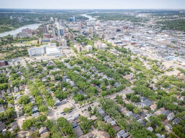 Şehir manzarası yukarıdan, içinden bir nehir akıyor. Şehir evler ve binalarla dolu ve caddelerde birçok araba var. Gökyüzü açık ve güneş parlıyor.