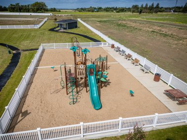 Çevresinde çit ve yeşil kaydırak olan bir oyun alanı. Arka planda banklar ve küçük bir bina var.