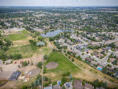 Park ve göleti olan bir yerleşim yeri manzarası. Evler dağıldı ve caddeye park etmiş birkaç araba var.