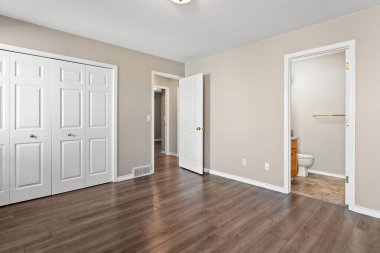 Beyaz kapısı ve beyaz dolabı olan büyük, boş bir yatak odası. Oda çıplak ve dağınık, ahşap zemin ve köşede tuvalet var.