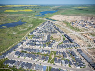 Evleri ve yolları olan bir yerleşim yeri manzarası. Evlerin çoğu beyaz ve caddeye park edilmiş birkaç araba var. Gökyüzü açık ve güneş parlıyor.