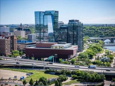 Şehir manzarası ve ortasında büyük bir bina. Bina ağaçlarla çevrili ve bir sürü penceresi var. Yolda arabalar ve arka planda bir kamyon var. Gökyüzü açık.