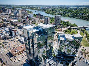 Şehir manzarası ve içinden bir nehir geçiyor. Binalar uzun ve yansıtıcıdır. Gökyüzü açık ve güneş parlıyor.