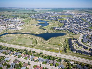 Ortasında göl olan yerleşim yeri manzarası. Evler dağılmış durumda ve yolda birkaç araba var. Gökyüzü açık ve güneş parlıyor.