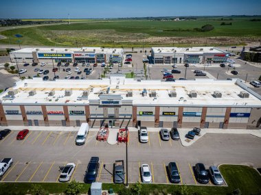 Bir sürü park yeri olan büyük bir alışveriş merkezi. Alışveriş merkezi araba ve kamyonlarla dolu ve etrafta dolaşan birkaç kişi var. Gökyüzü açık ve güneş parlıyor.