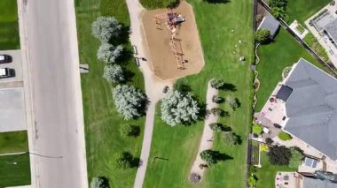 Ortasında park olan bir yerleşim bölgesinin sokak manzarası. Park ağaçlarla çevrili ve kaldırımı var. Arka planda birkaç ev var.