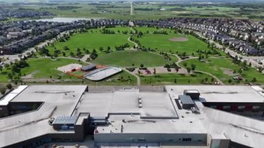 Basketbol sahası ve buz pateni sahası olan büyük bir park. Park evlerle çevrilidir. Gökyüzü mavi ve çimenler yeşil