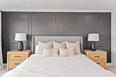 Beyaz çarşaflı ve yastıklı büyük yatak gri boyalı bir duvarın önünde duruyor. Oda birkaç lamba ve bir komodinle süslenmiş.