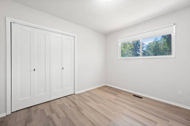 Penceresi ve dolabı olan beyaz bir yatak odası. Oda boş ve minimalist bir görünüşü var.