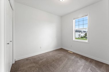 Penceresi ve kapısı olan bir yatak odası. Oda boş ve beyaz bir duvarı var. Pencere açık ve oda güneşle aydınlanıyor.