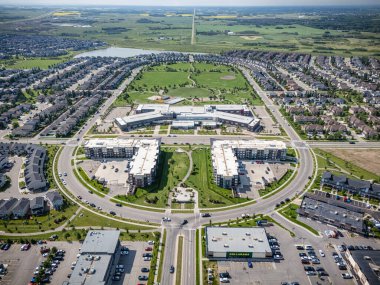 Merkezinde büyük yeşil bir park olan bir yerleşim yeri manzarası. Park, büyük bir alışveriş merkezi de dahil olmak üzere birkaç binayla çevrilidir. Cadde park halindeki arabalarla dolu.
