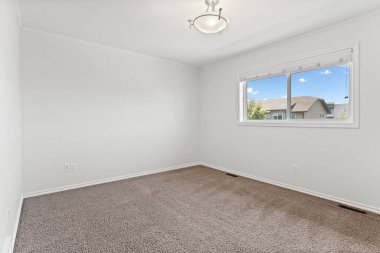 Büyük, penceresi ve aydınlatma armatürü olan boş bir oda. Oda çıplak ve dağınık, halı kaplı zemini ve beyaz bir duvarı var. Pencere içeri doğal ışık salıyor, parlak ve havadar bir atmosfer yaratıyor.