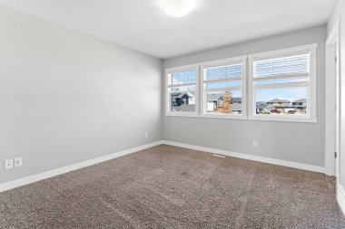 Beyaz bir duvarı ve beyaz halısı olan çok temiz ve boş bir yatak odası. Odanın üç penceresi var, biri açık.