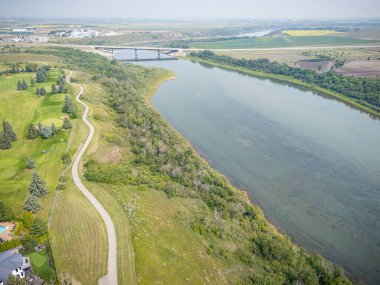 Bir köprüsü ve yanında uzanan bir yolu olan bir nehir. Yol ağaçlar ve otlarla dolu.