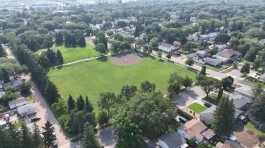 Arka planda beyzbol sahası ve birkaç ev olan bir park manzarası. Park ağaçlarla çevrili ve yeşil çimenli bir arazisi var.