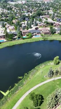 Arka planda ağaçlar ve gölet olan bir golf sahası manzarası. Gölet yeşil çimenlik bir alan ve bir patikayla çevrilidir. Ağaçlar uzun ve yayılmış. Huzurlu ve huzurlu bir atmosfer yaratıyorlar.