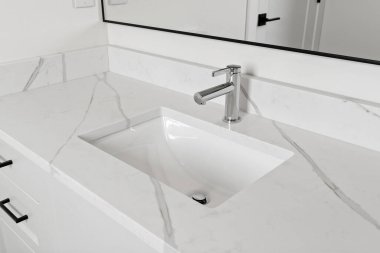 Üstünde gümüş musluk ve ayna olan bir lavabo. Lavabo beyaz, ayna siyah.