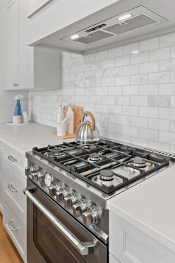 White kitchen with a stove top oven and a silver tea kettle on top of it. The stove has a clock on it and a few bottles and vases on the counter