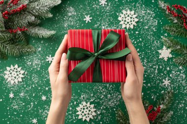 Yeni yıl konsepti. Kadın ellerinin kırmızı hediye kutusuna kurdele, kar tanesi süslemesi, kar ökse otu ve yeşil arka planda donmuş çam dalları veren ilk kişi.