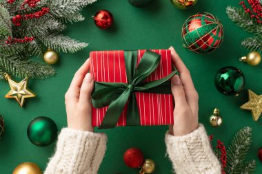 Noel konsepti. Örgü süveter giyen, kırmızı hediye kutusu taşıyan, altın sarısı takılar takan, ökseotu ve köknar dalları olan, yeşil arka planda bulunan ilk kişinin resmi.