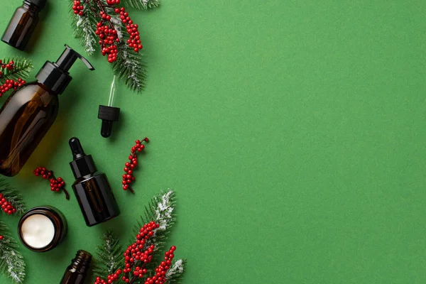 Winter skin care concept. Top view photo of cosmetic dispenser bottle cream jar dropper bottles spruce branches in frost and mistletoe berries on isolated green background with copyspace