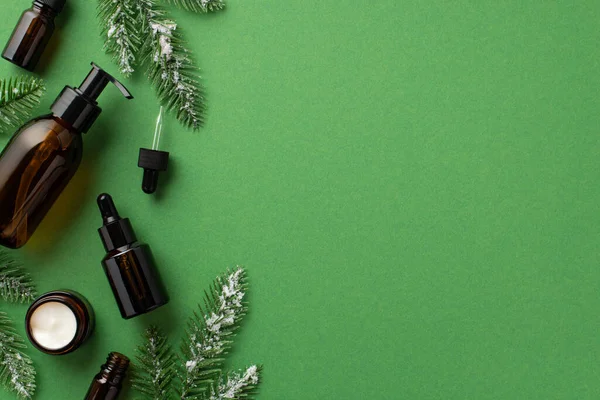 Winter season skin care concept. Top view photo of glass pump bottle cream jar dropper bottles and pine branches in hoarfrost on isolated green background with blank space