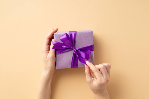 Christmas Eve concept. First person top view photo of female hands unpacking lilac giftbox with violet ribbon bow on isolated pastel beige background