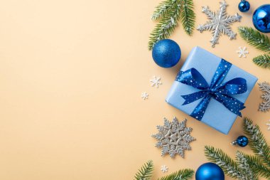 Noel arifesi kavramı. Mavi süs eşyalarının en üstteki fotoğrafı büyük hediye kutusu. Kurdele yay parlıyor kar tanesi süslemeleri dondaki çam dalları ve izole edilmiş pastel bej arka planda boş alan üzerine konfeti.