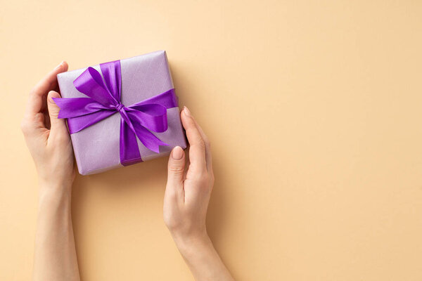 New Year concept. First person top view photo of female hands holding lilac giftbox with violet ribbon bow on isolated pastel beige background with empty space