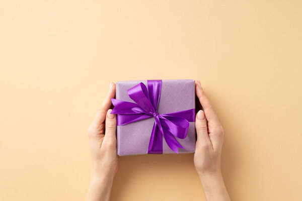 New Year concept. First person top view photo of female hands giving purple giftbox with violet ribbon bow on isolated pastel beige background with blank space