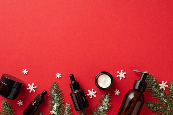 Winter skin care cosmetics concept. Top view photo of glass dispenser bottle cream jars dropper bottles spruce branches in hoarfrost and snowflakes on isolated red background with copyspace