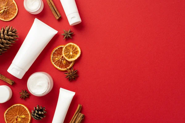 Organic skin care cosmetics concept. Top view photo of white tubes cream jars pine cones anise dried orange slices and cinnamon sticks on isolated red background with copyspace