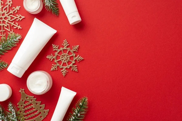 Winter skin care concept. Top view photo of white cosmetic bottles christmas decorations golden snowflake and fir ornaments on isolated red background with empty space