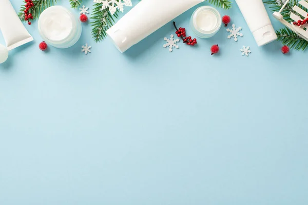 Winter season skin care concept. Top view photo of white tubes cream jars pine branches christmas ornaments mistletoe berries and snowflakes on isolated pastel blue background with empty space