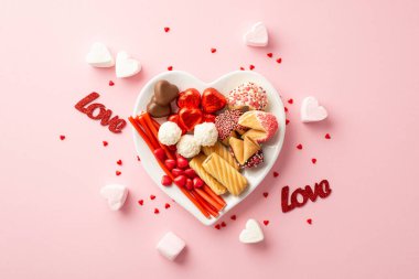 Sevgililer Günü konsepti. Kalp şeklindeki tabağın en üstteki fotoğrafı şekerli kurabiyeler çikolatalı jöle şekerler ve izole edilmiş pastel arkaplandaki yazıtlar.