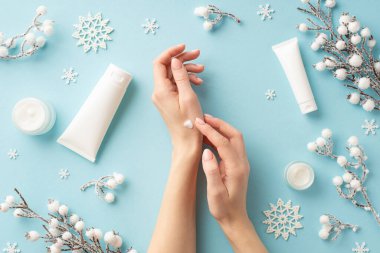Kışın cilt bakımı. Kadın ellerinin kozmetik kavanozlarına krem sürerken, kar taneleri ve izole edilmiş pastel mavi arka planda don ve bitki dalları sürerken çekilmiş ilk resmi.