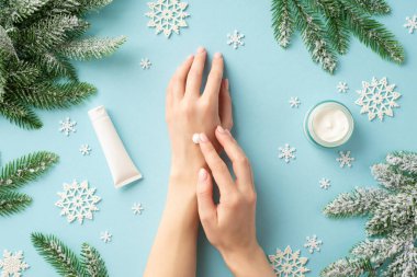 Kışın cilt bakımı. Ellerine krem sürerken çekilmiş ilk fotoğrafta kozmetik kavanoz kar taneleri ve çam dalları, izole açık mavi arka planda, kopyalanmış