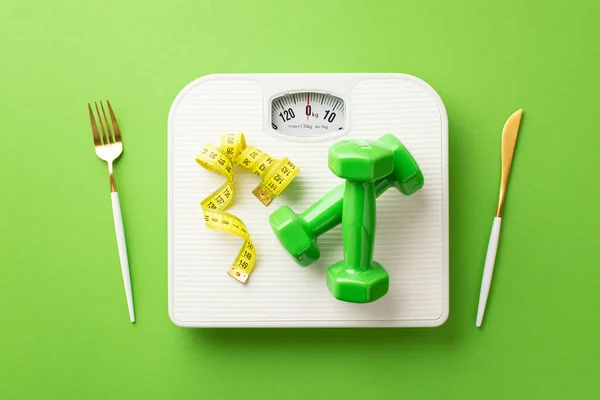 Weight loss concept. Top view photo of cutlery fork knife dumbbells tape measure and scales on isolated green background