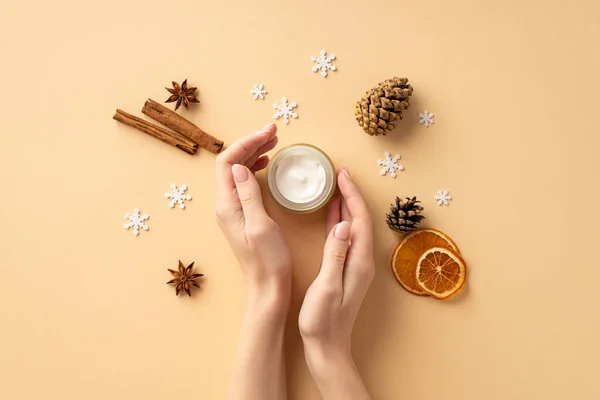 Winter season skin care concept. First person top view photo of female hands cream jar pine cones dried orange slices cinnamon sticks anise and snowflakes on isolated pastel beige background