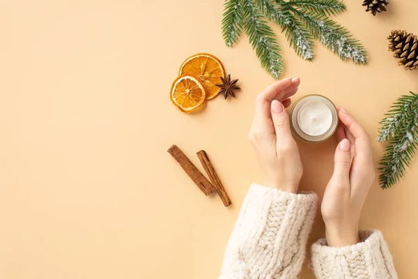Winter skin care concept. First person top view photo of female hands in sweater cream jar fir branches dried orange slices cinnamon sticks pine cone on isolated pastel beige background with copyspace