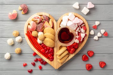 Sevgililer Günü konsepti. Tahta kalp şeklindeki servis tepsisinin en üst görüntüsü. Çikolatalı jöleli kurabiyeler ve gri ahşap masa arkasındaki cam bardak içeceği.