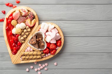 Sevgililer Günü konsepti. Tahta kalp şeklindeki servis tepsisinin en üst görüntüsü. Çikolata tabağı, jöle şekerler ve gri ahşap masa zemininde kurabiyeler, boş alan.