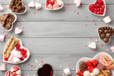 Sevgililer Günü konsepti. Kalp şeklindeki fincan tabakları, çikolatalı jöleli kurabiyeler, içki bardakları ve konfetilerin gri tahta masa altyapısı ve ortada boş bir alan.