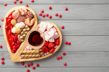 Sevgililer Günü konsepti. Ahşap kalp şeklindeki servis tepsisinin en üstteki fotoğrafı şekerleme bardakları ve kurabiyelerle doldurulmuş gri ahşap masa zemininde fotokopi
