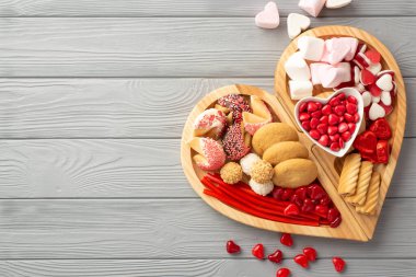 Sevgililer Günü kutlaması konsepti. Tahta kalp şekilli servis tepsisinin en üst görüntüsü. Tatlı tabağı, çikolata jöleli şekerler ve kurabiyeler. Gri ahşap masa zemininde.