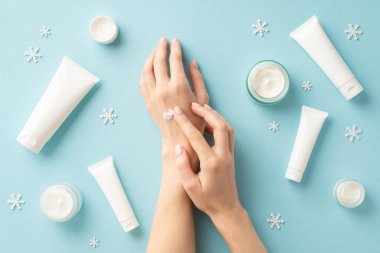 Kış cilt ürünleri konsepti. Kızın ellerine krem sürerken çekilmiş ilk resmi etiketsiz kavanozlar ve kopyalanmış açık mavi arka planda kar taneleri.