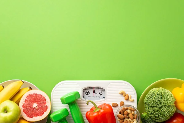 Slimming concept. Top view photo of plates with fruits and vegetables grapefruit apple bananas pepper cabbage cucumber nuts dumbbells and scales on isolated green background with copyspace